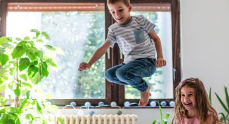 Gas Dekoratives Bild Zwei Kinder springen spielerisch in die Luft auf einem Sofa. Im Hintergrund befindet sich ein Fenster mit einer grünen Pflanze davor.