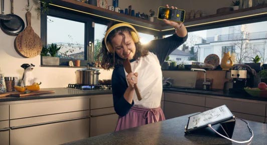 Kombi-Bonus-100-Euro Dekoratives Bild Ein Mädchen steht in der Küche und hört über Kopfhörer Musik. Sie benutzt einen Kochlöffel als Mikrofon und hält tanzend ihr Smartphone in die Lüfte.