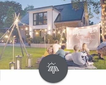 solar Eine Familie sitzt gemütlich auf dem Rasen auf einer Decke und schaut einen Film auf einer Projektionsleinwand. Sie sind umgeben von dekorativen warmweißen Lichterketten. Im Hintergrund ein modernes Haus mit einer Solaranlage auf dem Dach.